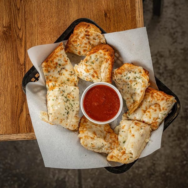 A basket of cheesy breadsticks with herbs and a side of marinara sauce on a wooden table.