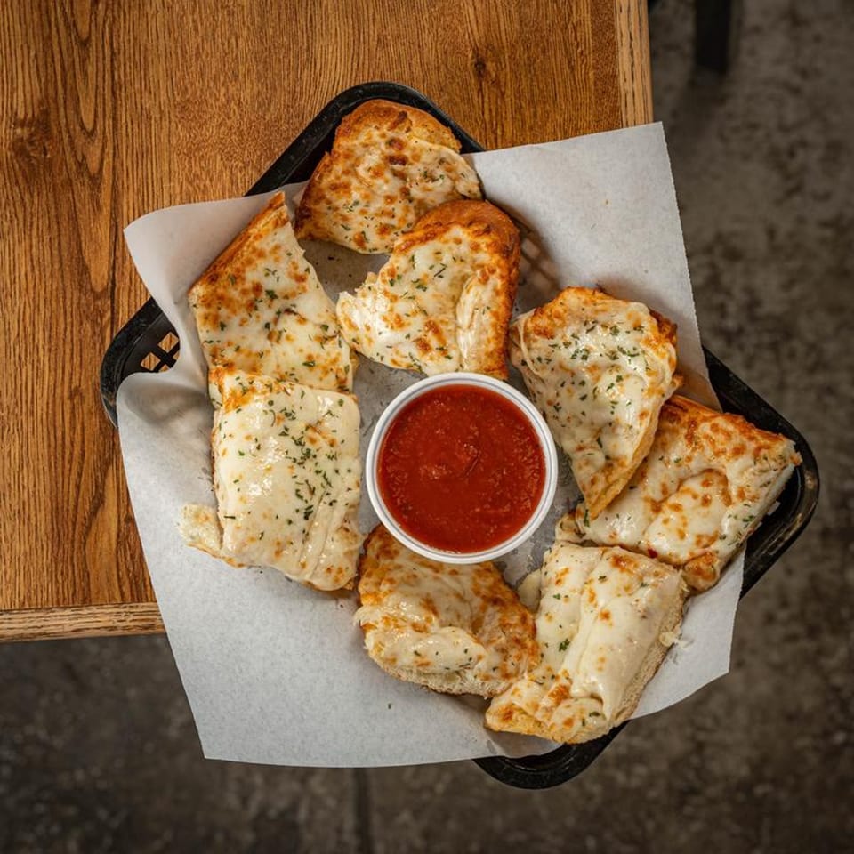 A basket of cheesy breadsticks with herbs and a side of marinara sauce on a wooden table.