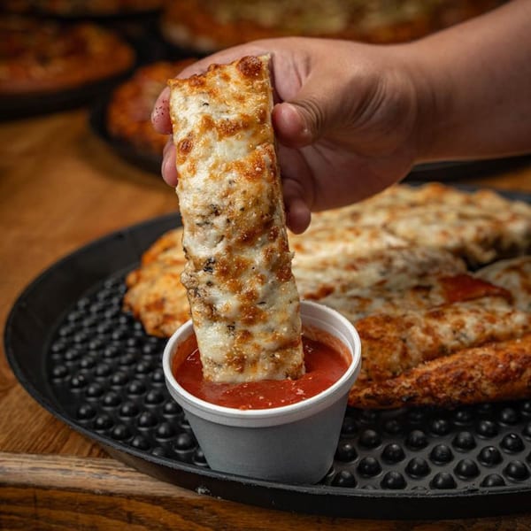 A hand dips a cheesy breadstick into a small cup of red marinara sauce.