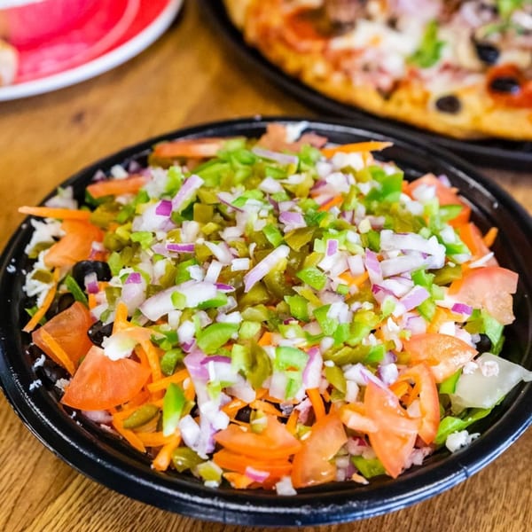A fresh salad with diced tomatoes, onions, peppers, and olives, next to a blurred pizza.